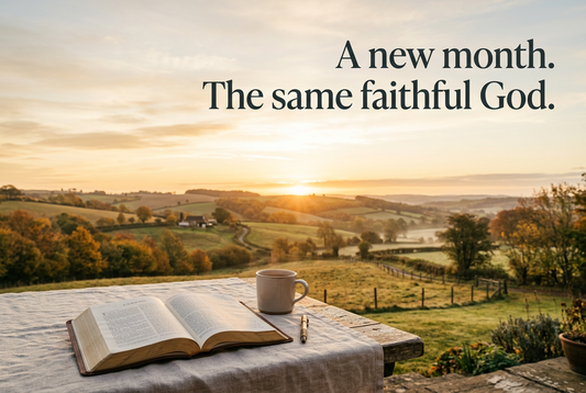 Open Bible and cup on a linen-covered table overlooking countryside at sunrise, with the words “A new month. The same faithful God.”